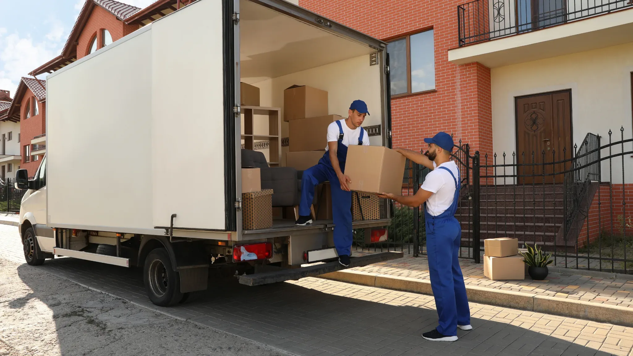 A mover in a blue cap and blue coveralls with white straps stands inside the open back of a white box truck, passing a large