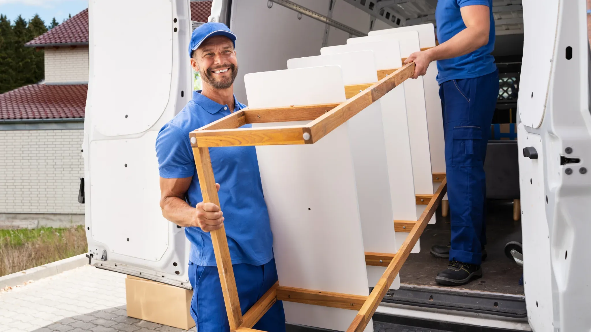 A smiling mover in a blue cap and blue coveralls over a white t-shirt looks at the camera while carrying a tan cardboard box